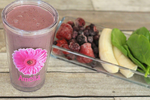 Kid friendly green smoothie in a personalized glass with spinach, Greek yogurt, frozen blueberries, blackberries and strawberries and berries, banana in a glass bowl on a wood table