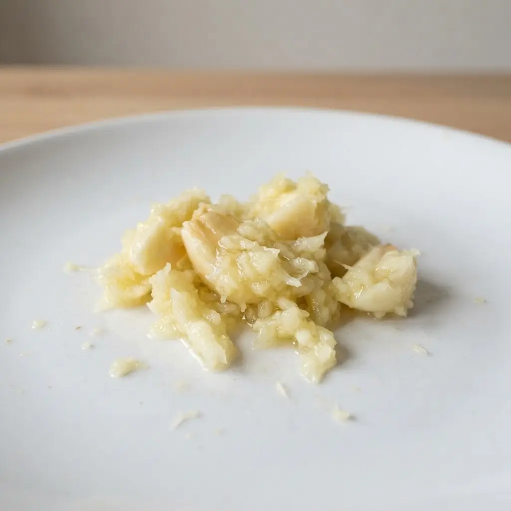 A macro view of crushed garlic cloves shows moist surfaces and fine pieces on a white plate.